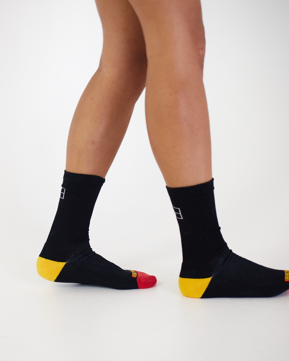 Clothing The Gaps. Long black socks with white Aboriginal flag outline on back. Yellow on heel of socks. Red on toes of socks. Clothing The Gaps written above red section just going over toes.